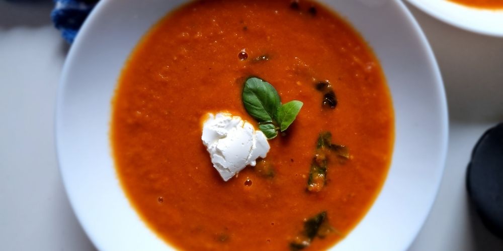 Tomato soup in a white plate with sour cream on top and a leaf of fresh basil.