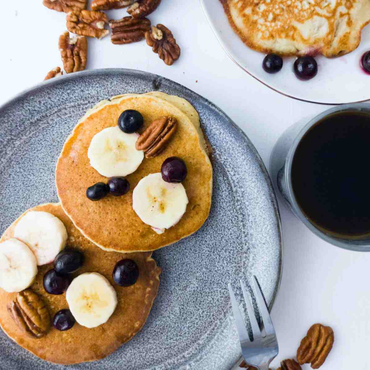 Pancakes on a plate with sliced banana, blueberries, and pecans on top.