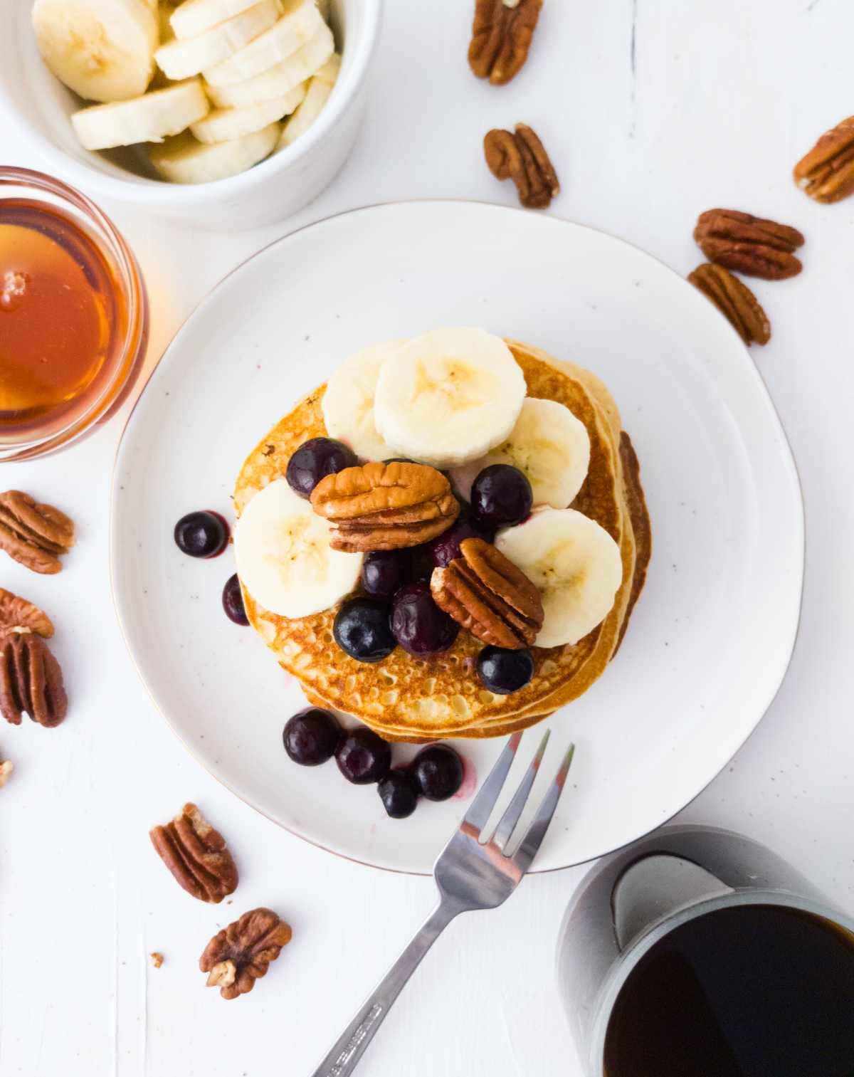 Gluten-free banana sourdough pancakes on a white plate with bananas, blueberries, and pecans on top.