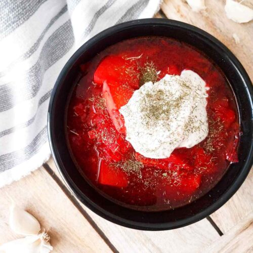 Traditional borscht in a black bowl with a spoon of sour cream on the top against of a wooden surface.