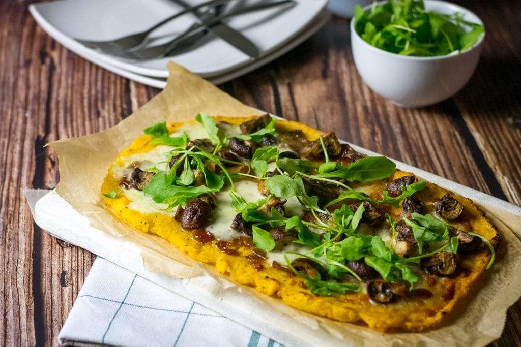 Polenta pizza on a wooden table with a plate and forks in the background.