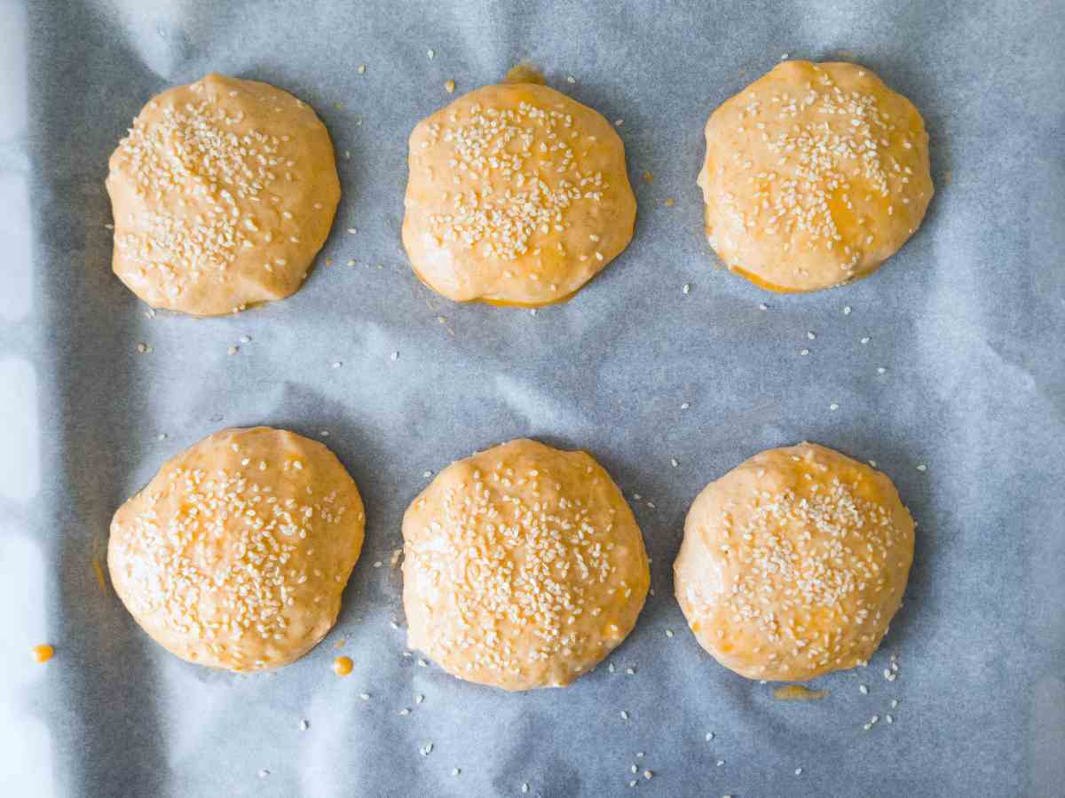 Hamburger buns brushed with egg and topped with sesame seeds on a baking sheet lined with parchment paper.
