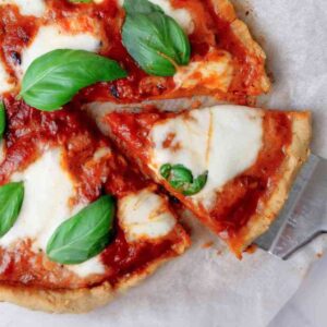Cassava pizza with a slice cut out and held by a metal spatula on a piece of white parchment paper.