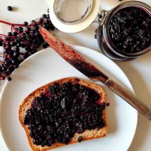 A slice of bread on a white plate covered with elderberry jam with a knife on the side of the plate and the jam in the background.