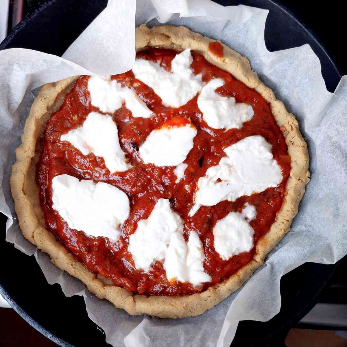 Par-baked cassava pizza crust with raw toppings on a white parchment paper against a black background.