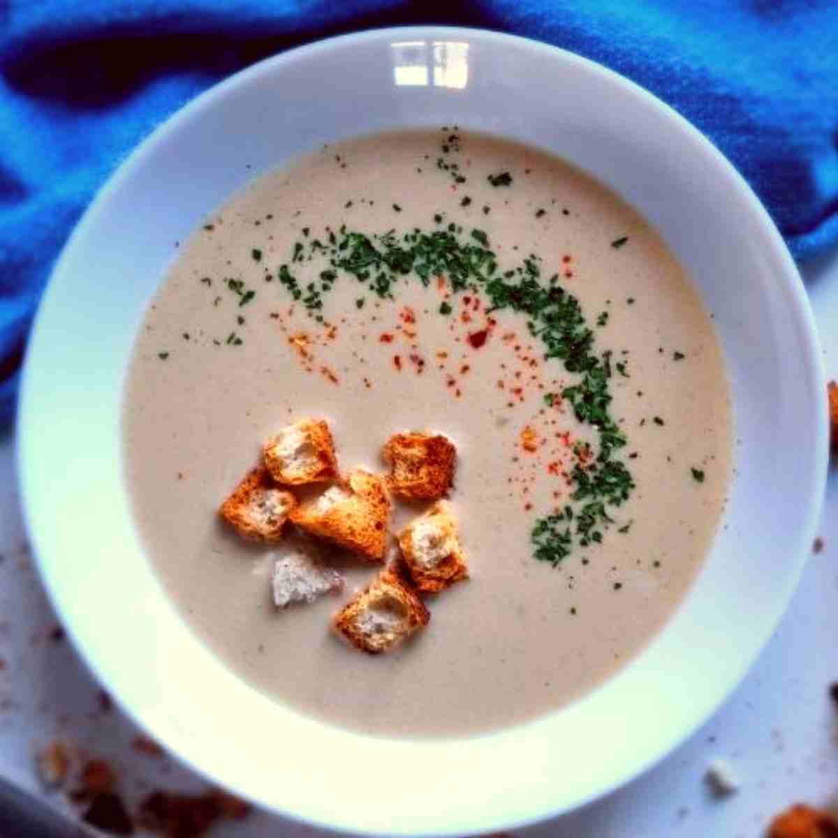 Garlic soup with croutons and green herbs in a white plate on a white table.