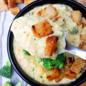 A bowl of Broccoli cheddar soup with carrots with a spoon and croutons.