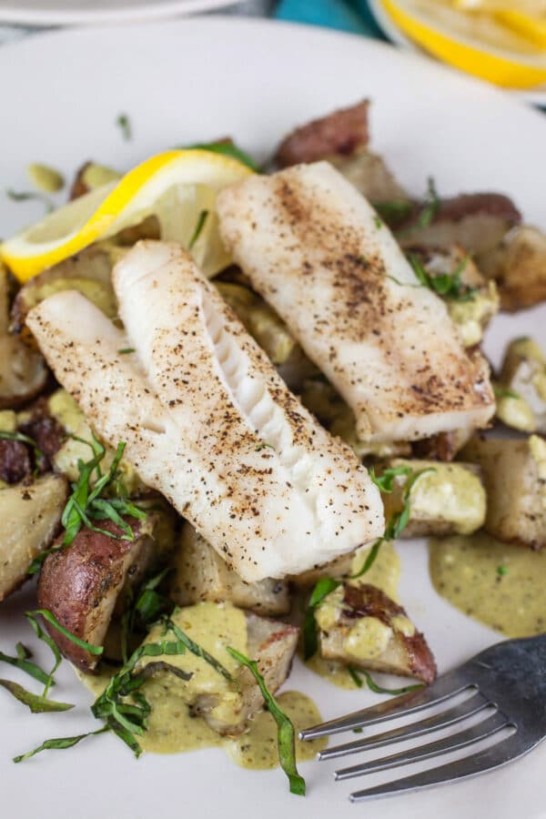 Two pieces of cod on a white plate together with potatoes, lemon, and greens.