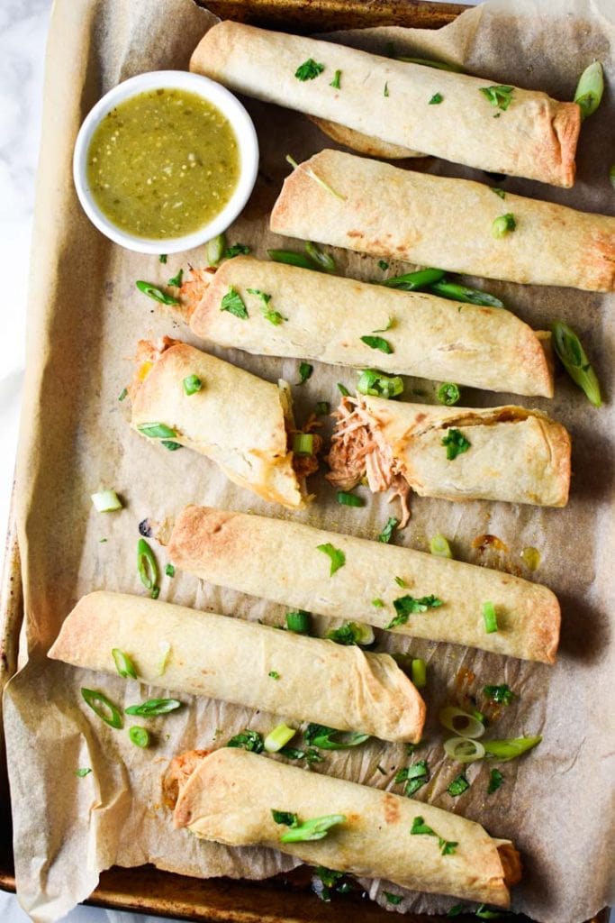 Baked creamy chicken taquitos on a tray with sauce on the side.