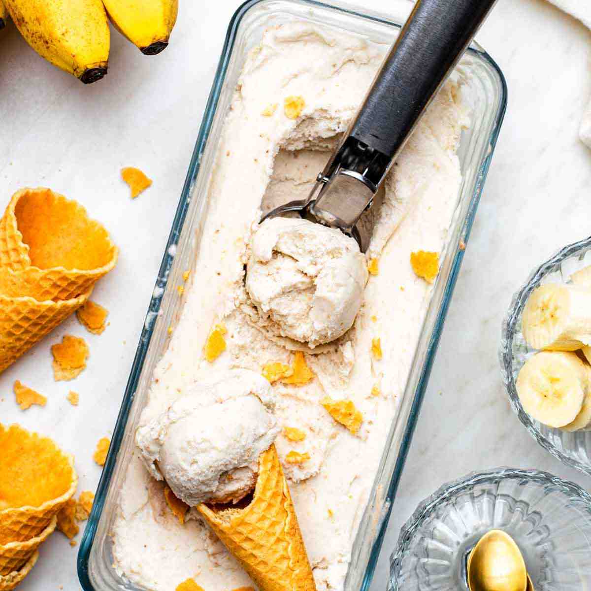 Banana ice cream in a container with an ice cream scooper and a cone in it,