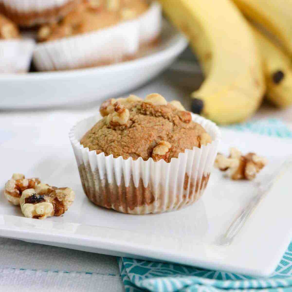 Banana oat muffin on a white plate with walnuts on the sides and a banana in the background.