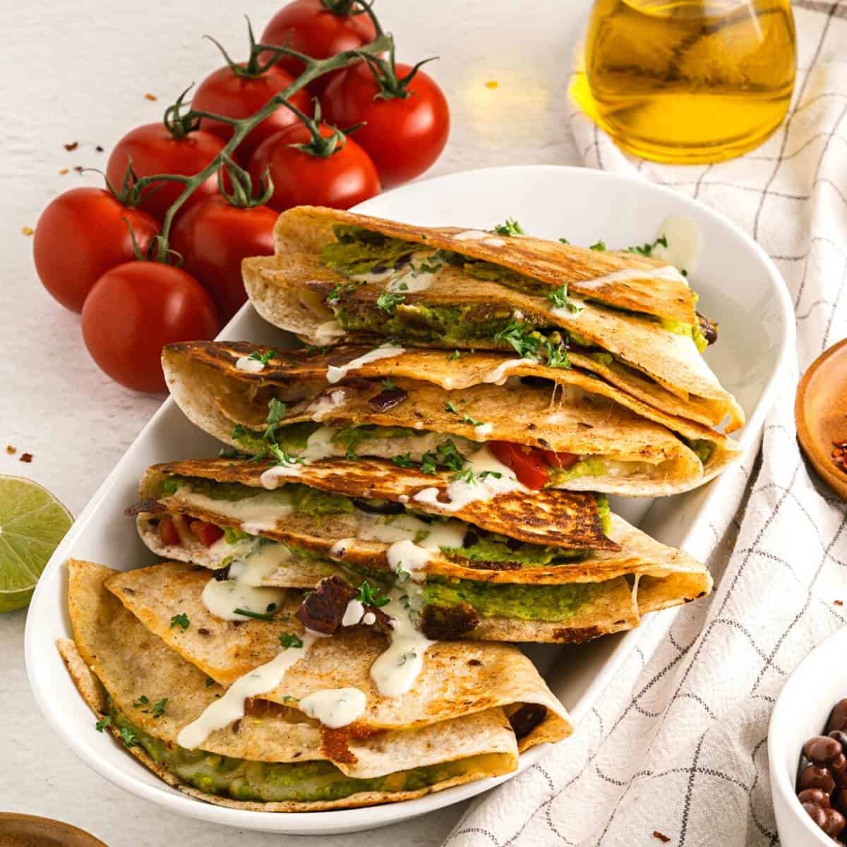 Guacamole quesadillas in a stack on a white dish with tomatoes in the background.