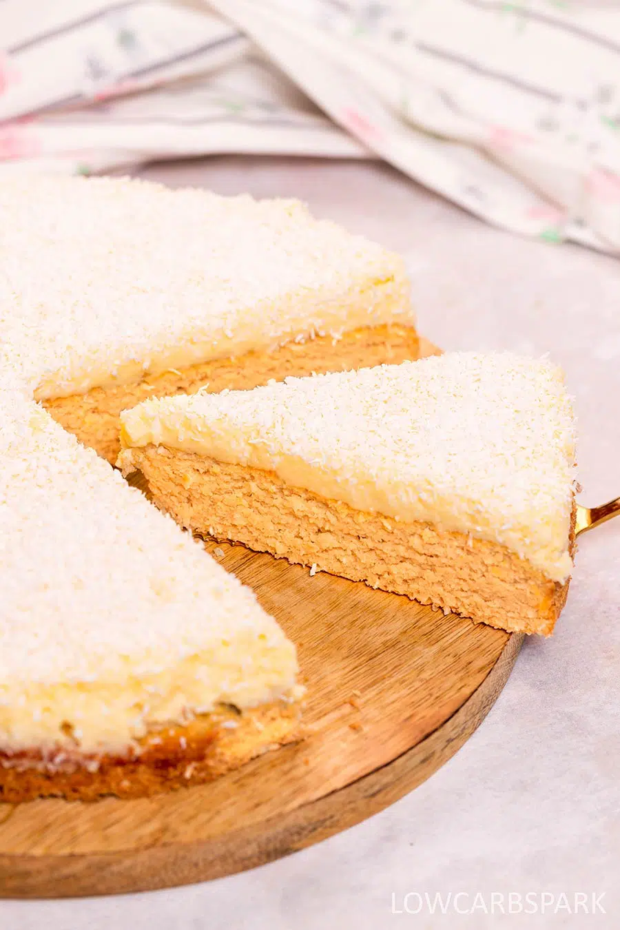 Coconut cake on a wooden plate with a slice being taken out with a metal spatula.