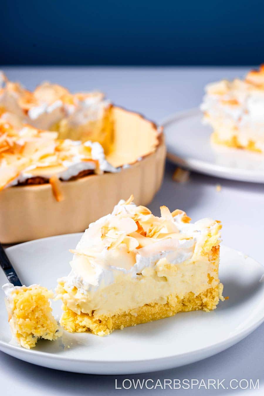 Coconut cream pie on a white plate with the rest of the pie in the background.