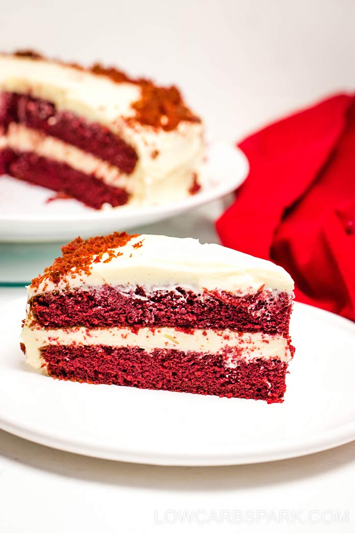 Red velvet cake slice on a white plate with the rest of the cake in the background.