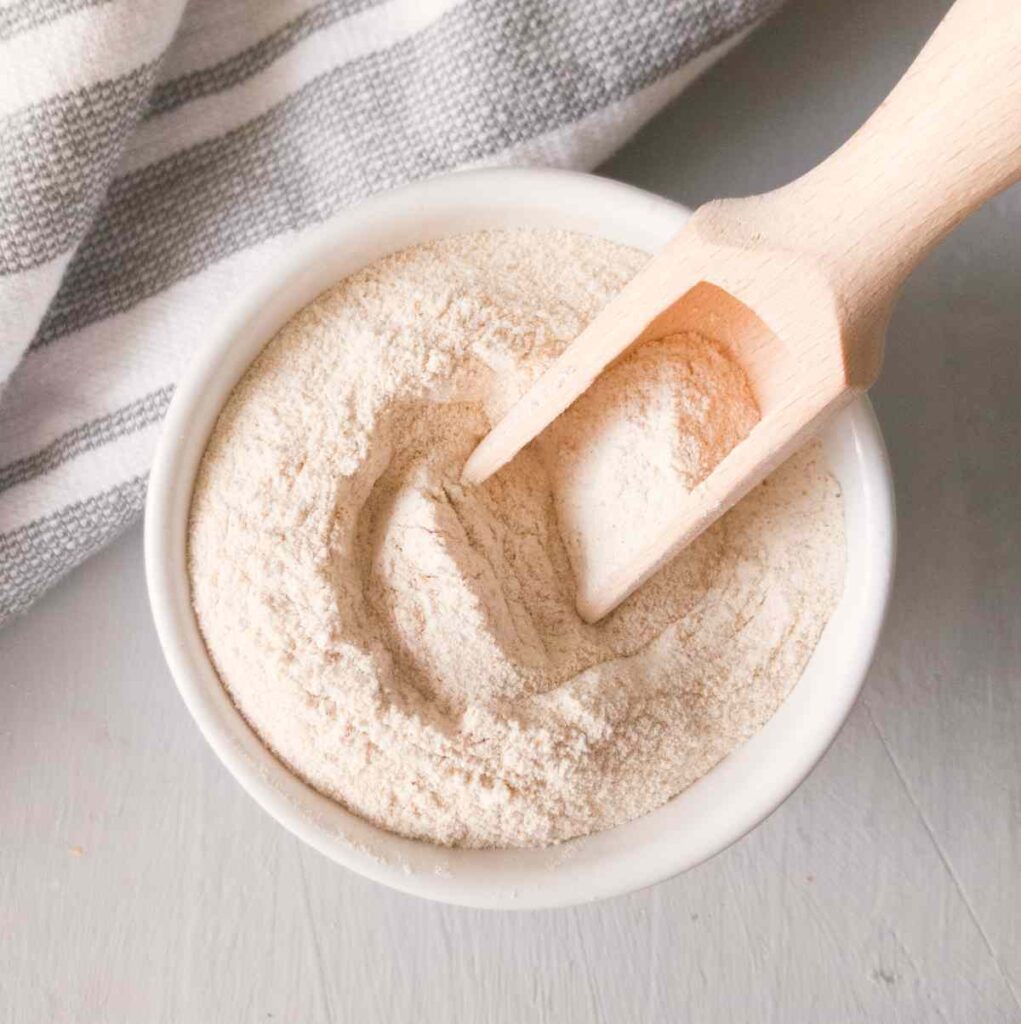 Buckwheat flour in a white bowl with a wooden spoon in it on a gray surface with a tea towel in the background.