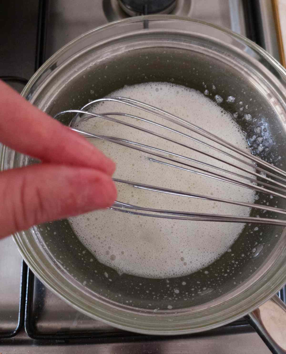 A metal saucepan with egg whites and a whisk on the stove with fingers over it.