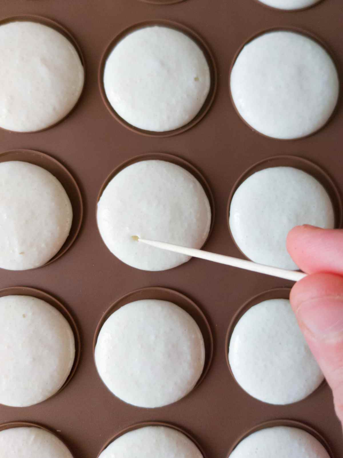 Piped macaron shells on a silicone mat with a toothpick popping bubbles.