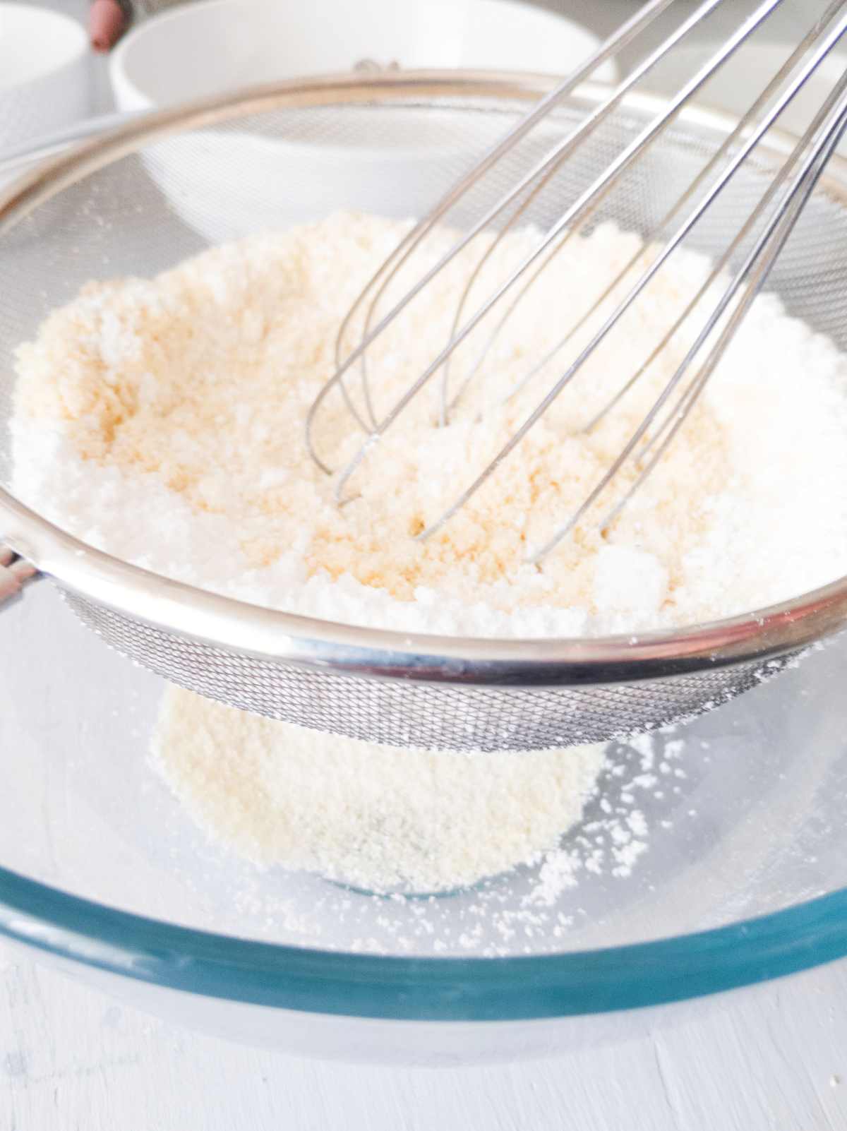 Sieving coconut flour and powdered sugar over a glass bowl with a whisk.