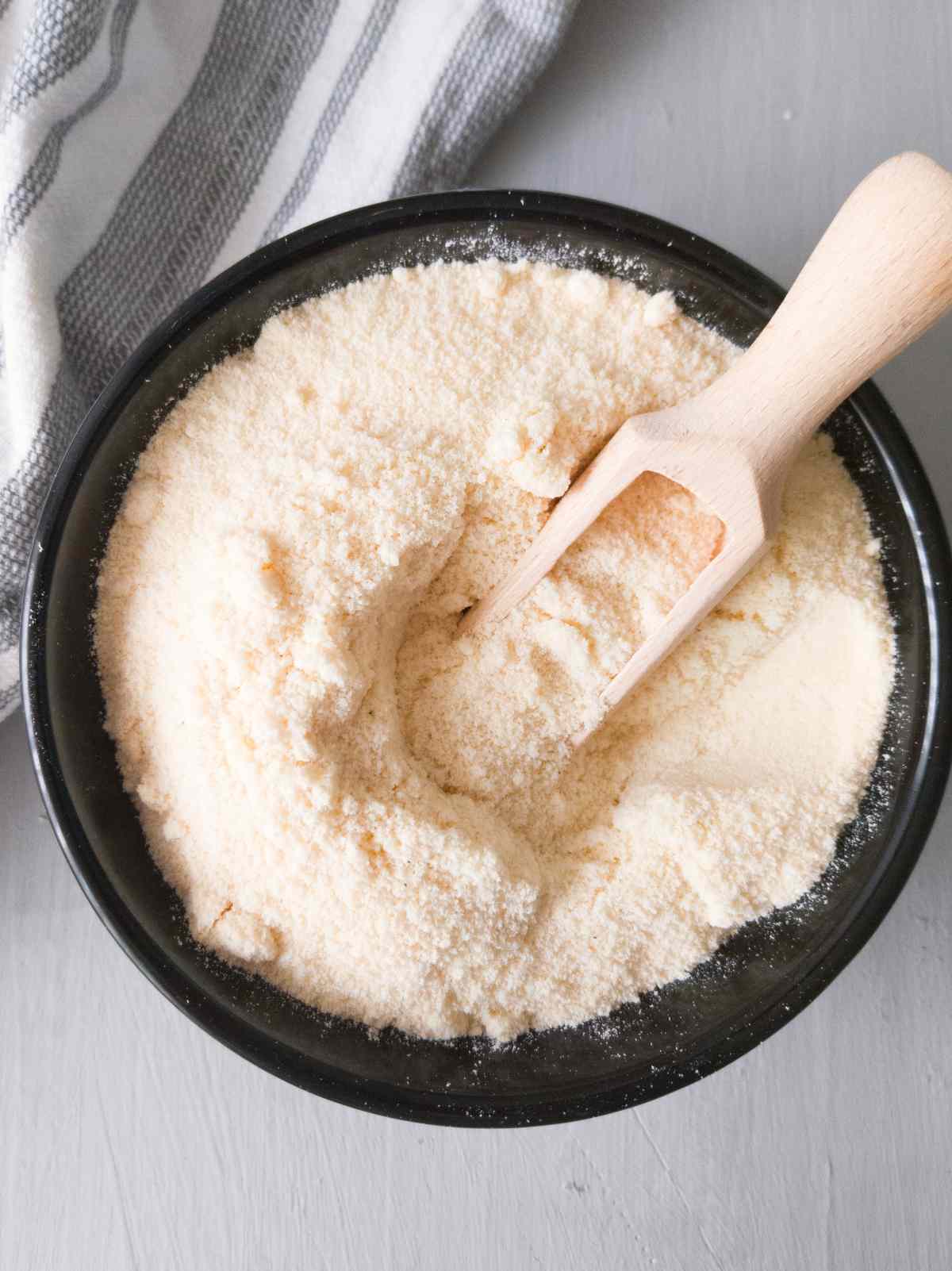 Coconut flour in a black bowl with a wooden spoon in it on a gray surface.