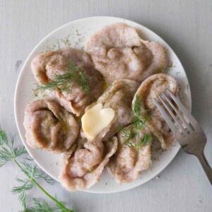 Gluten-free pelmeni on a round white plate with dill next to it and a fork on it.