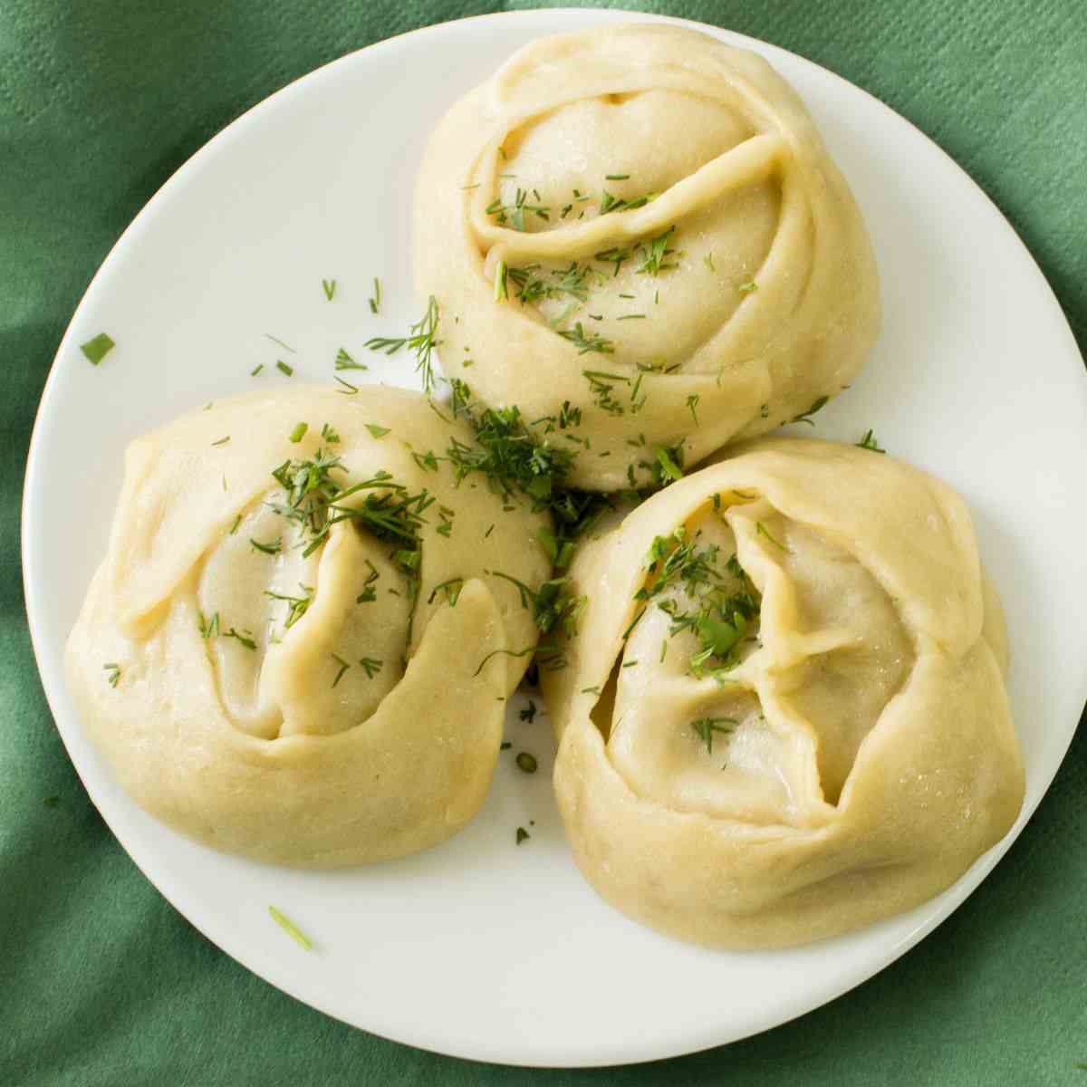 Manti on a white plate with a green background and chopped green onion sprinkled on top.