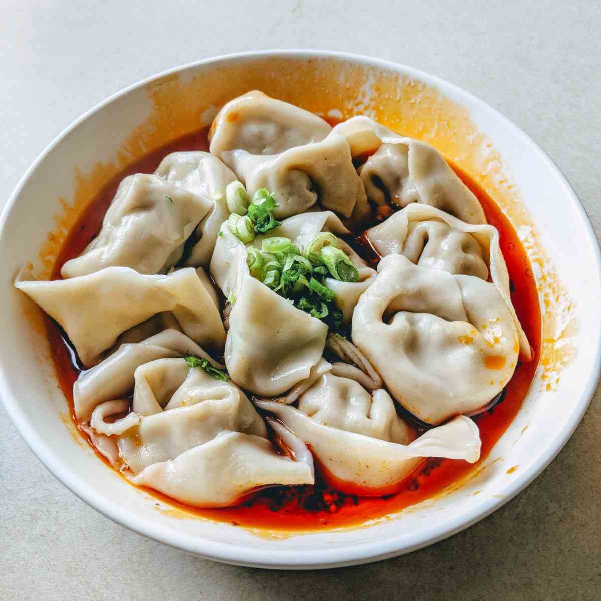 Chinese wonton dumplings in a bowl with red sauce and green onion on the top.