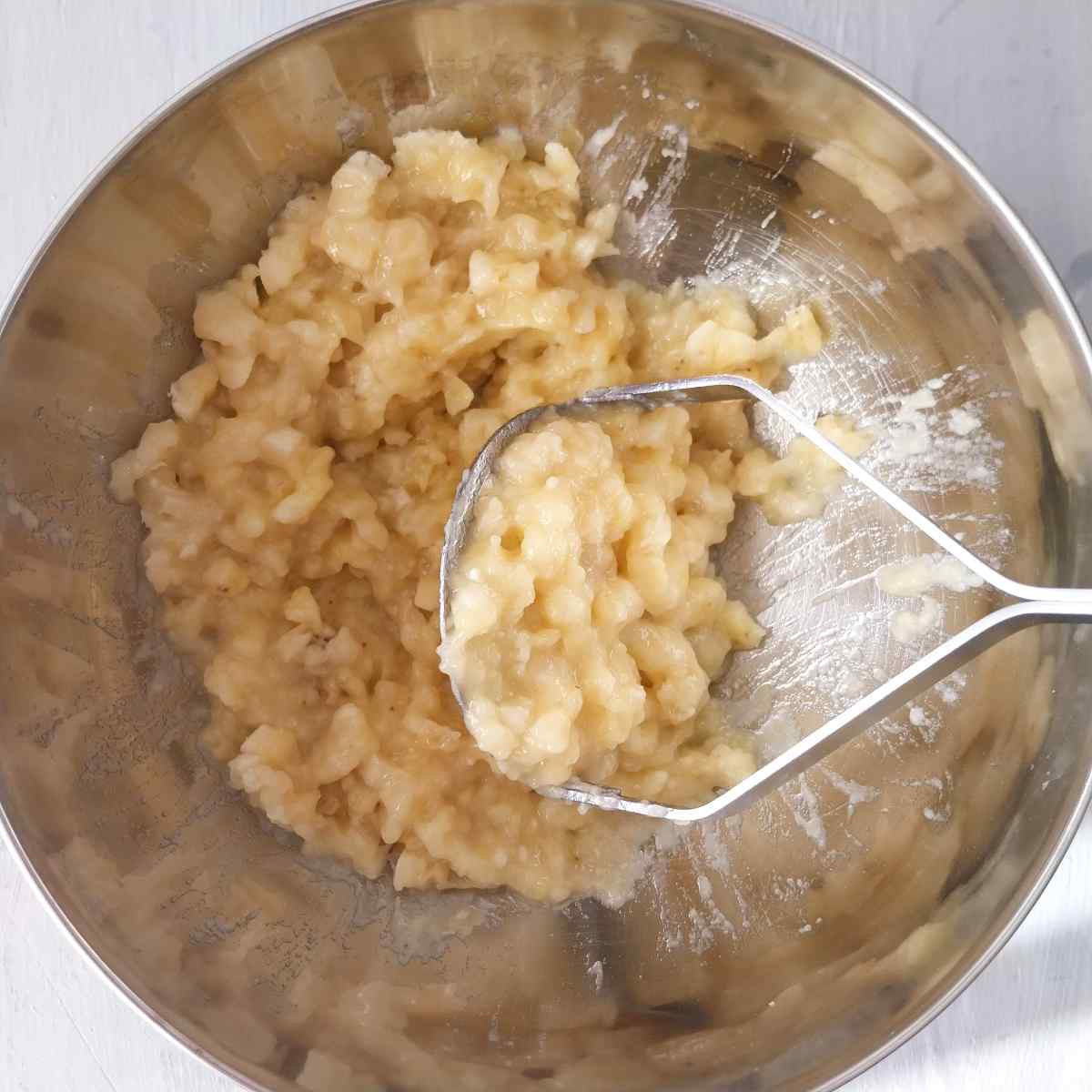 Mashed bananas with a masher in a metal bowl.