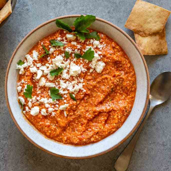 Red pepper dip in a small bowl with cheese and greens on the top. 