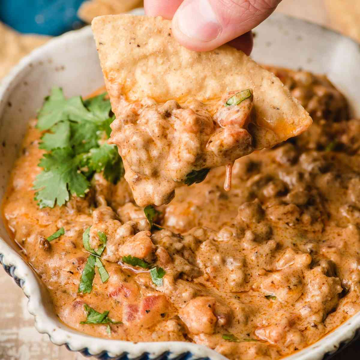 Beef queso dip in a bowl with a tortilla chip being dipped into it by a hand.