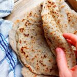 A flatbread being rolled by a hand on a wooden surface.