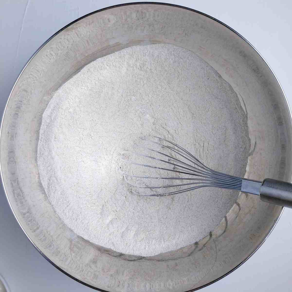 A metal mixing bowl with flour and a whisk in it.