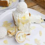 A slice of Raffello cheesecake on a white plate with Raffaello candy next to it and almond flakes sprinkled around.