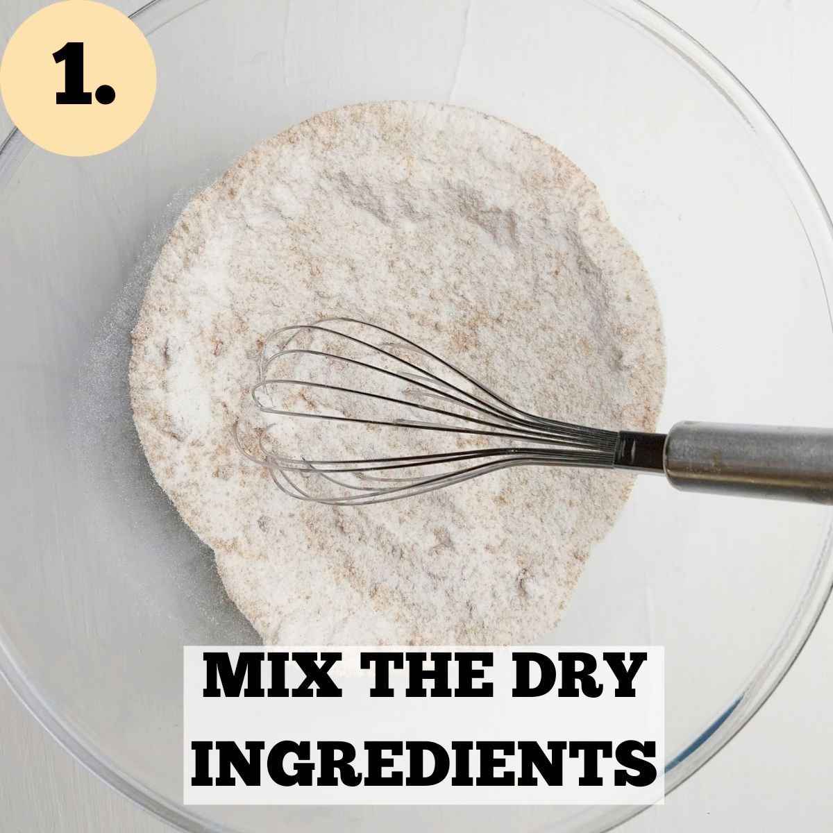 Dry ingredients in a glass mixing bowl with a whisk in it against a white surface.