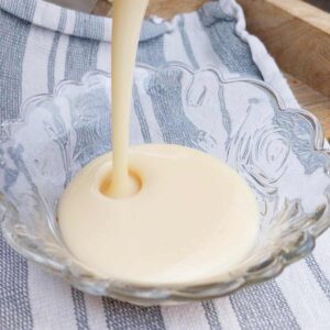 Pouring light creamy condensed milk into a small glass bowl sitting on a kitchen towel.