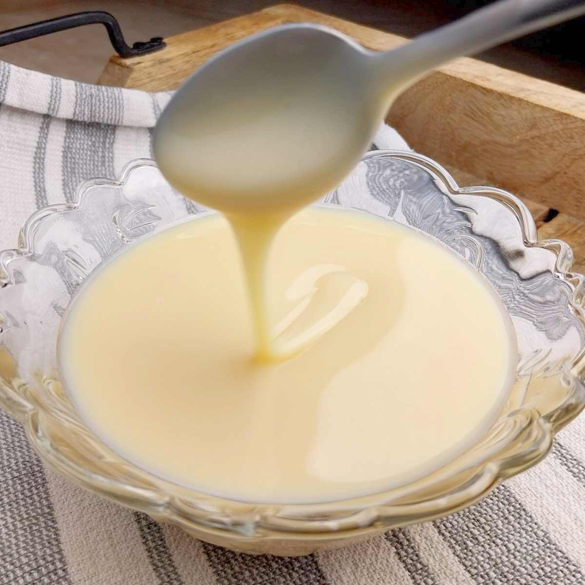 Sweetened condensed milk in a small glass bowl scooped with a tea spoon, sitting on a kitchen towel.