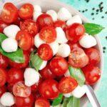 Cherry tomatoes with mozzarella balls and fresh basil in a large bowl with a spoon in it against green background.