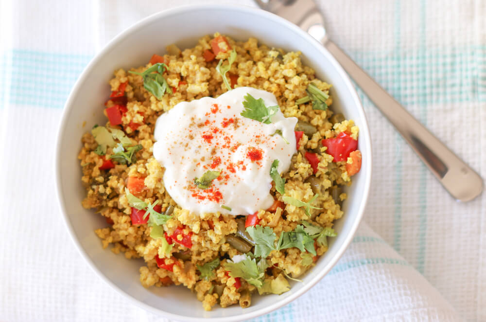 Millet stir fry in a white bowl with sour cream in the middle, with a spoon on the side on a white kitchen towel.