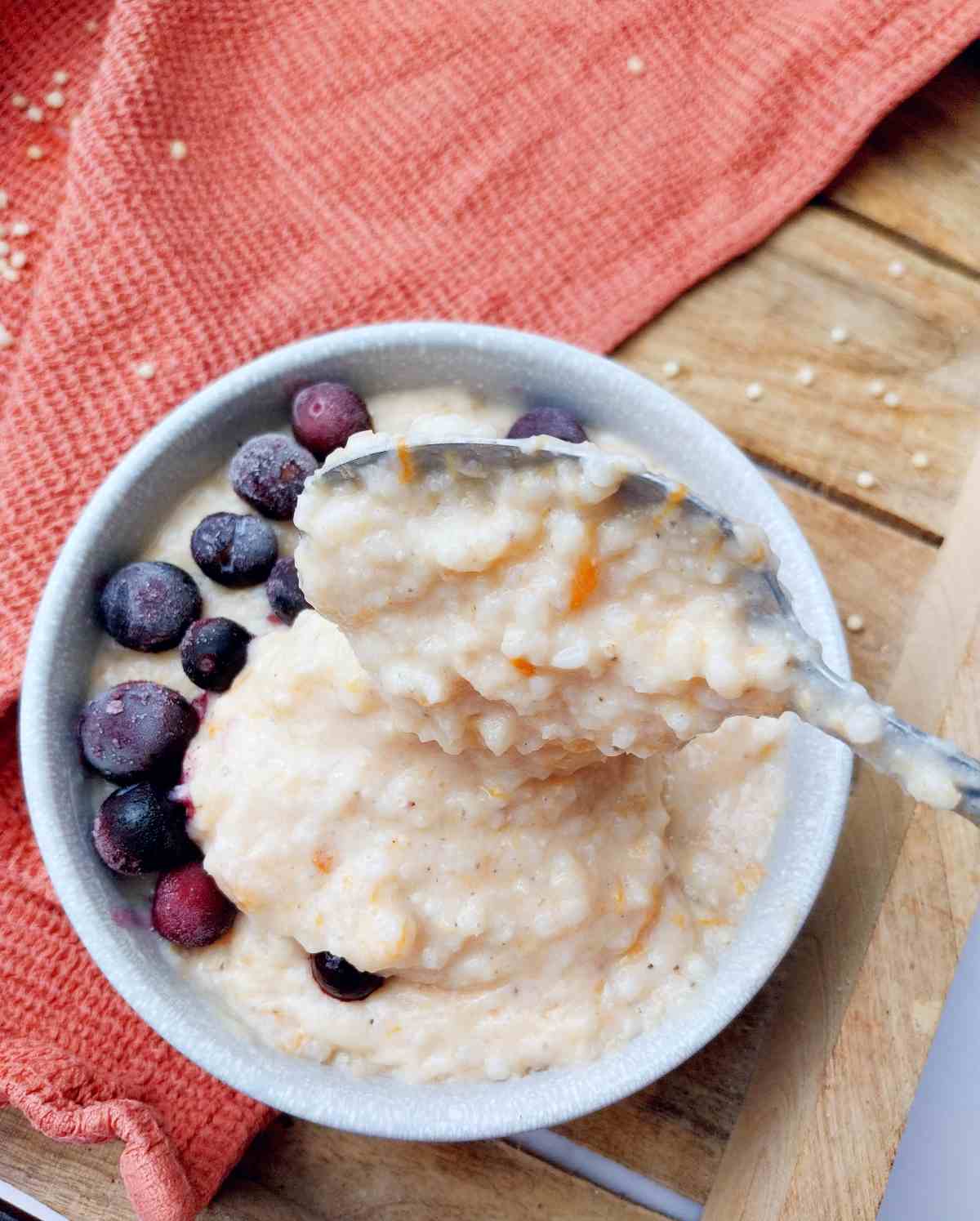 Sorghum porridge in a light colored bowl with frozen blueberries and a spoon in it on a wooden surface with a kitchen towel in the background.