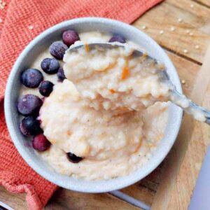 Sorghum porridge in a light colored bowl with frozen blueberries and a spoon in it on a wooden surface with a kitchen towel in the background.