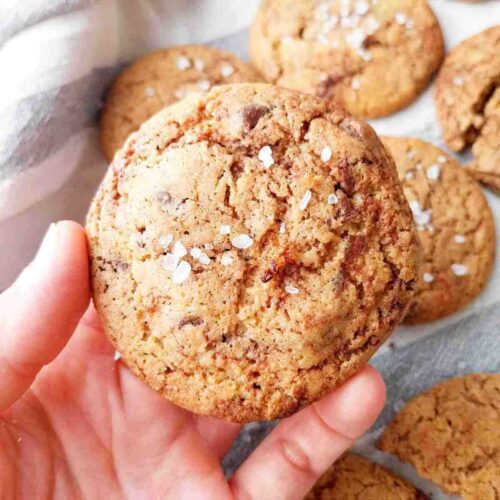 A millet flour cookie with flaky salt on top held in a hand with the rest of the cookies in the background on a kitchen towel.