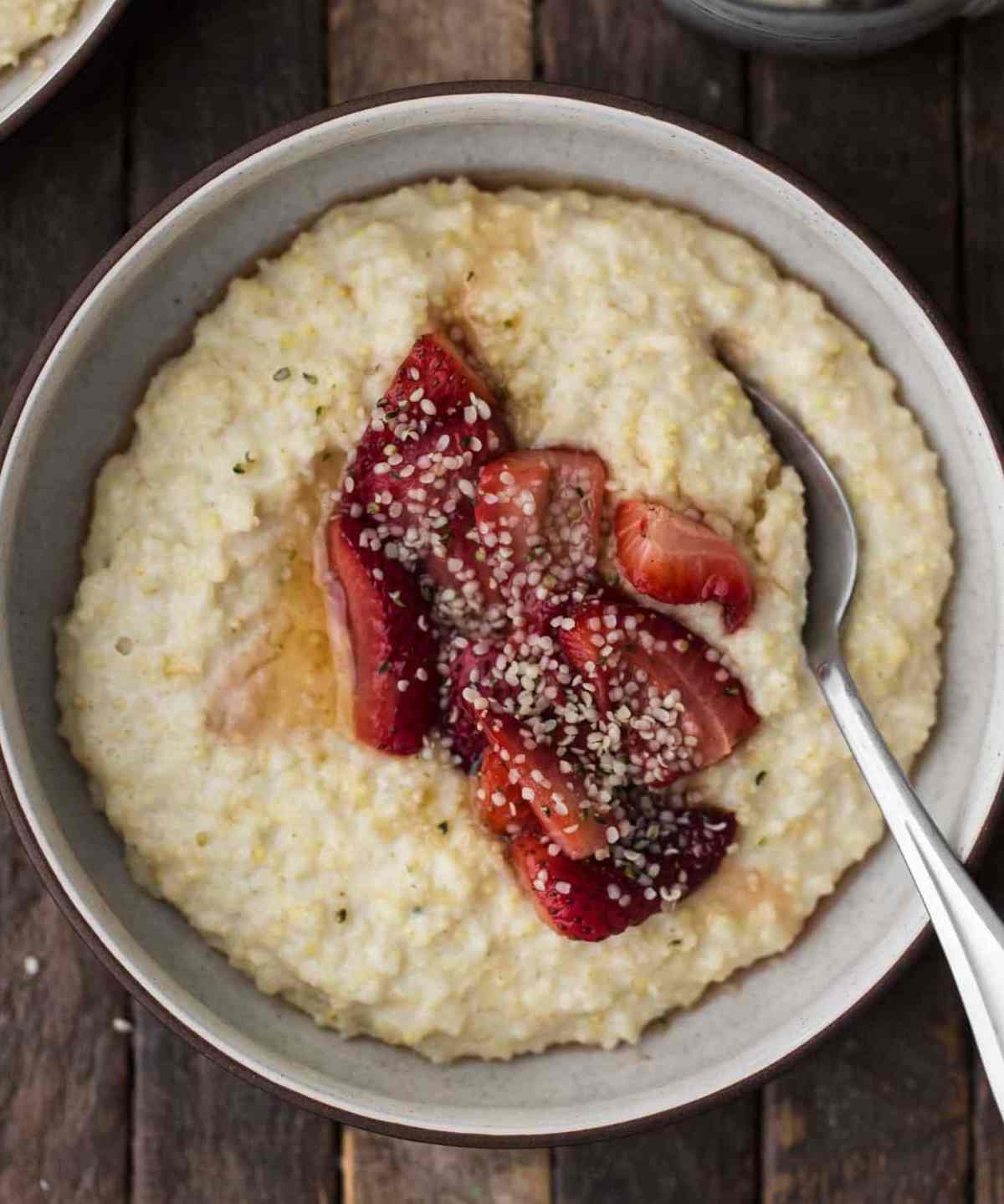 Millet porridge in a white bowl with strawberries on top and a spoon sticking out of the bowl on a wooden surface.