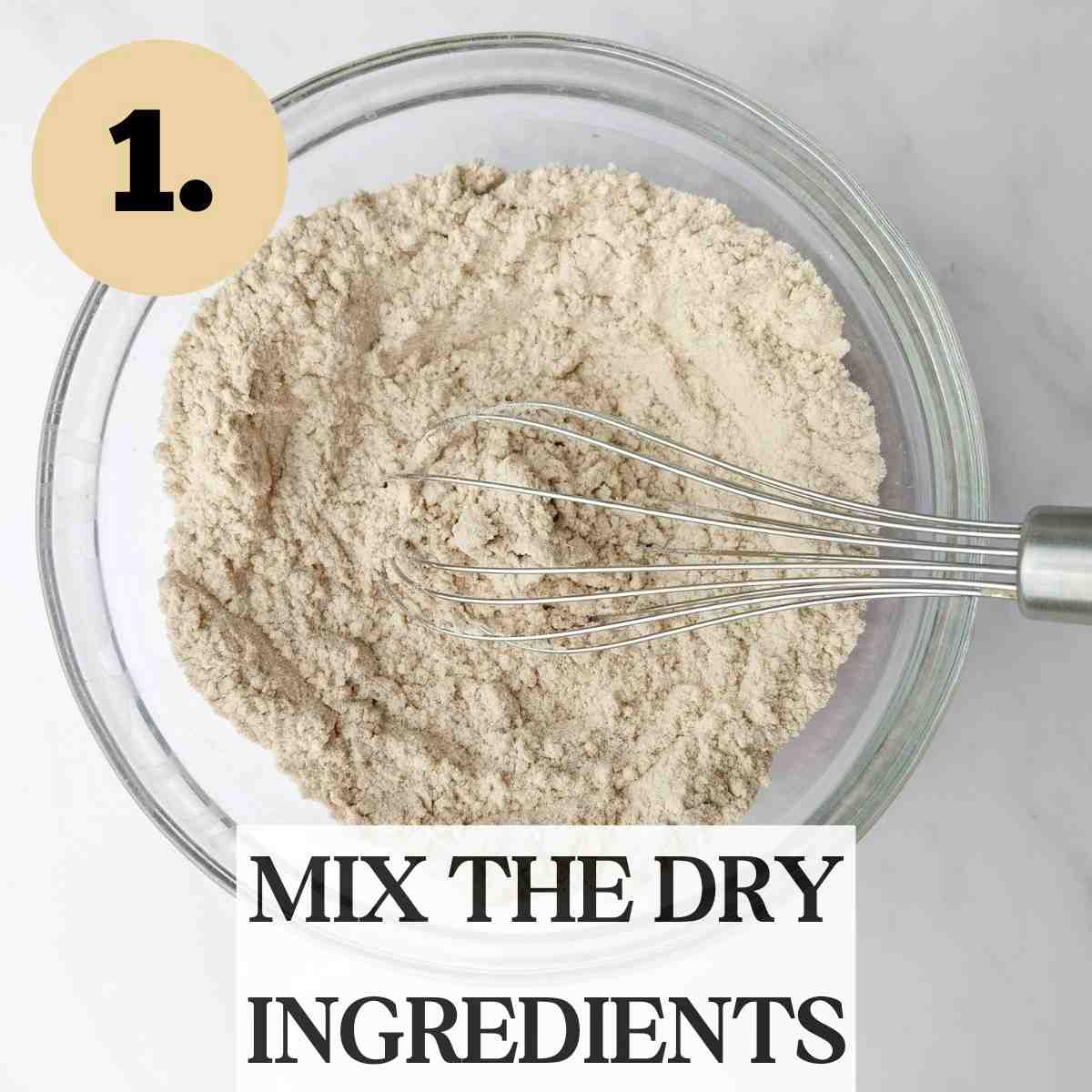 Dry sorghum cookie ingredients in a glass bowl with a metal whisk in it on a white surface.