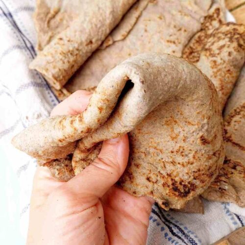 Ragi roti bent and held in a hand with the rest of the rotis in the background on a light kitchen towel.