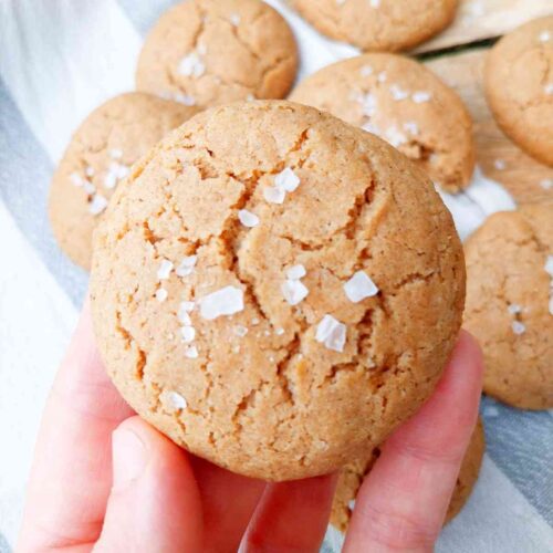 Sorghum cookie with flaky salt on top held in a hand with the rest of the cookies in the background on a kitchen towel.