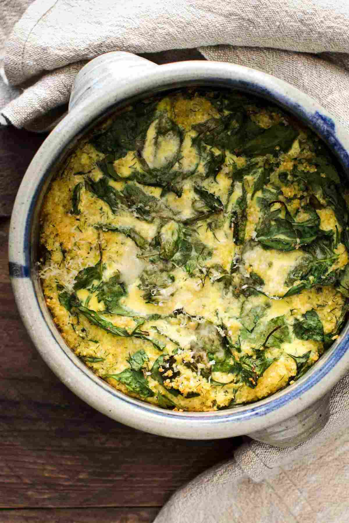 A deep baking dish with egg millet meal with spinach on top on a dark surface with a light kitchen towel wrapped around it.